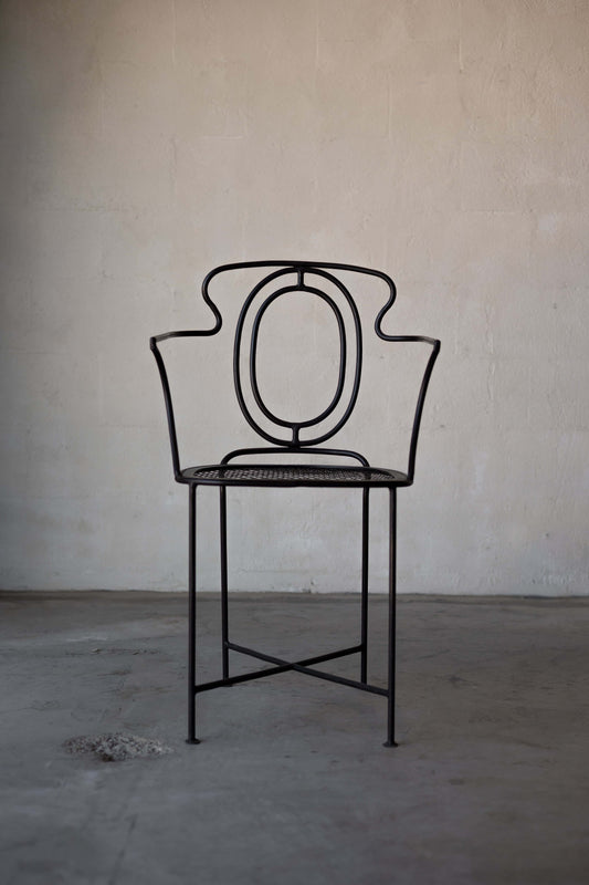 A black, wrought iron dining chair with art-deco design featuring a rounded, open backrest with curved accents. The seat is made of a perforated metal mesh, and the legs are slender with a crisscross support structure.