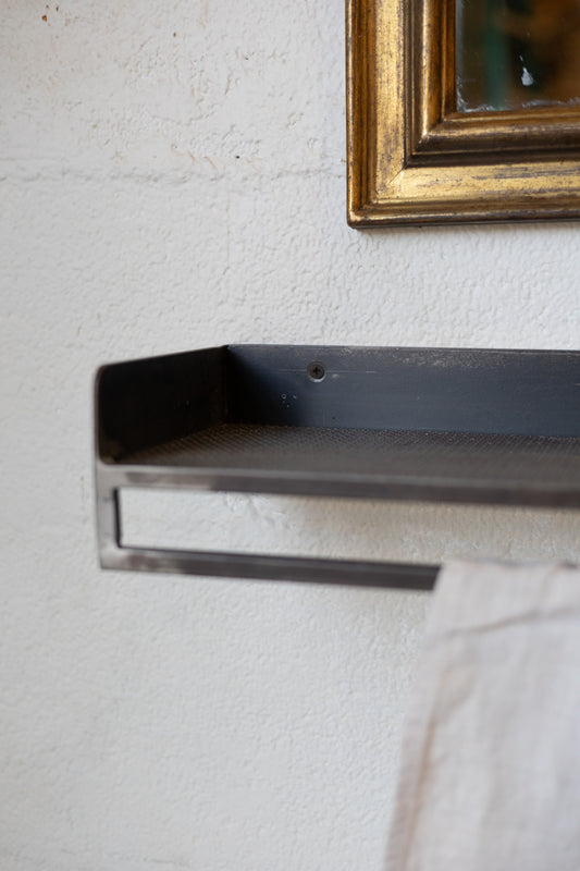 Metal shelf mounted on a textured wall with a gold-framed mirror above and linen hanging on rack.