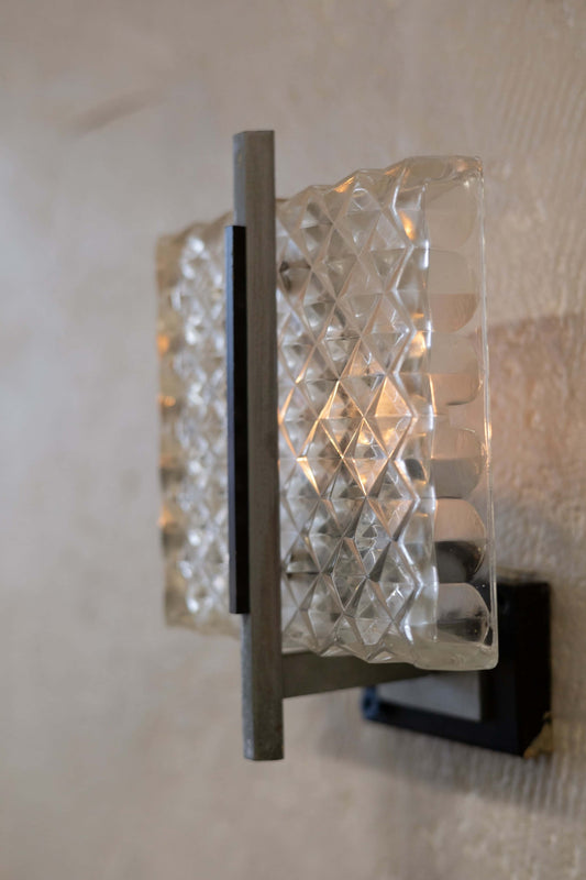 Vintage wall sconce featuring a prism glass shade with a diamond pattern, emitting a warm glow. The sconce is supported by a sleek metal frame with a minimalist design, mounted on a black metal base.