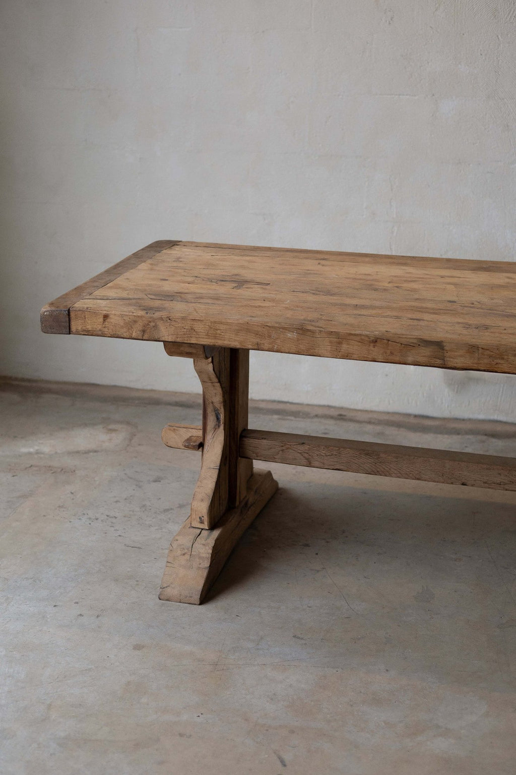View of wooden dining table's smooth top and natural, warm-toned grain. Features trestle base, curved leg, and horizontal support beam.