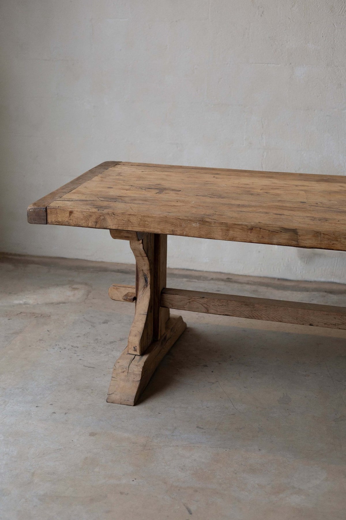 View of wooden dining table's smooth top and natural, warm-toned grain. Features trestle base, curved leg, and horizontal support beam.