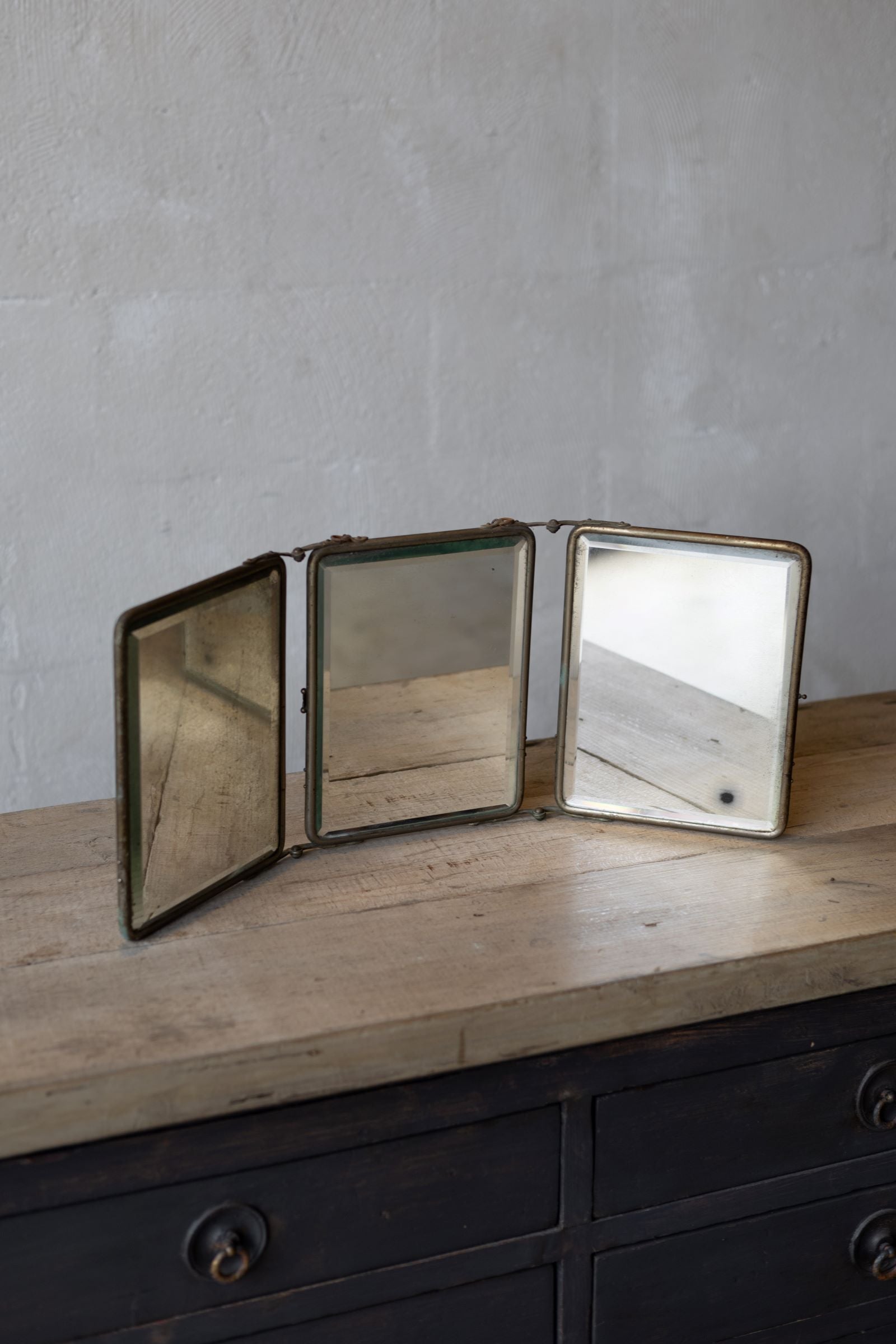Three-panel vintage mirror on a wooden surface with a gray wall background