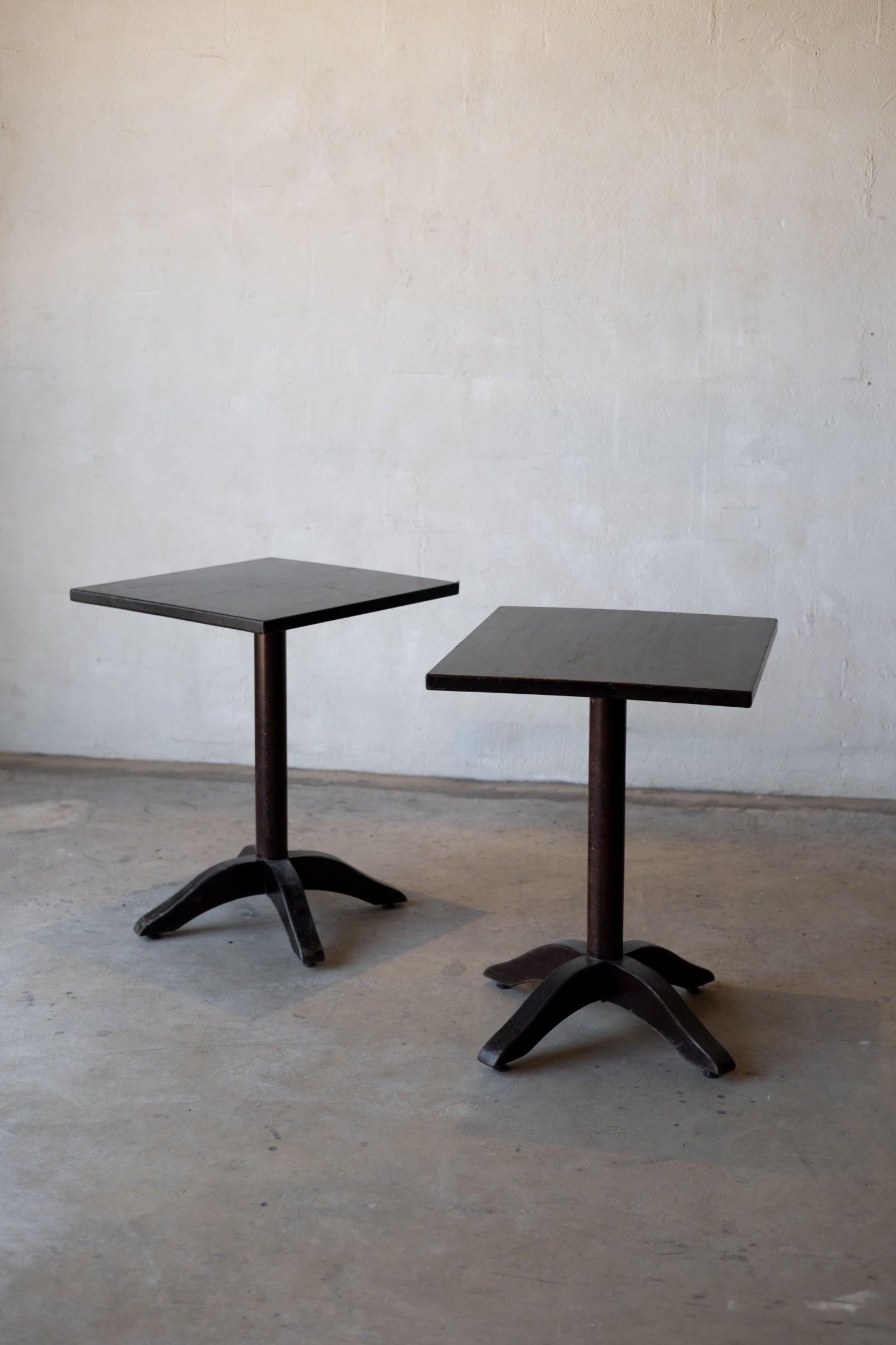 Two dark brown tables with pedestal bases on a plain floor against a white wall.