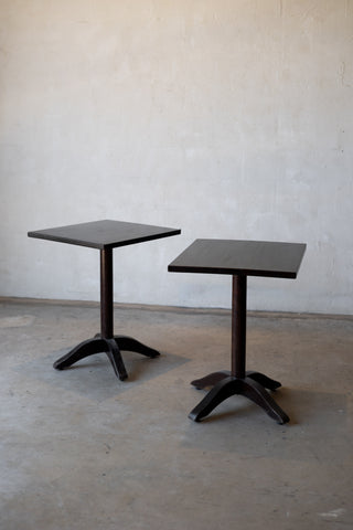Two dark brown tables with pedestal bases on a plain floor against a white wall.