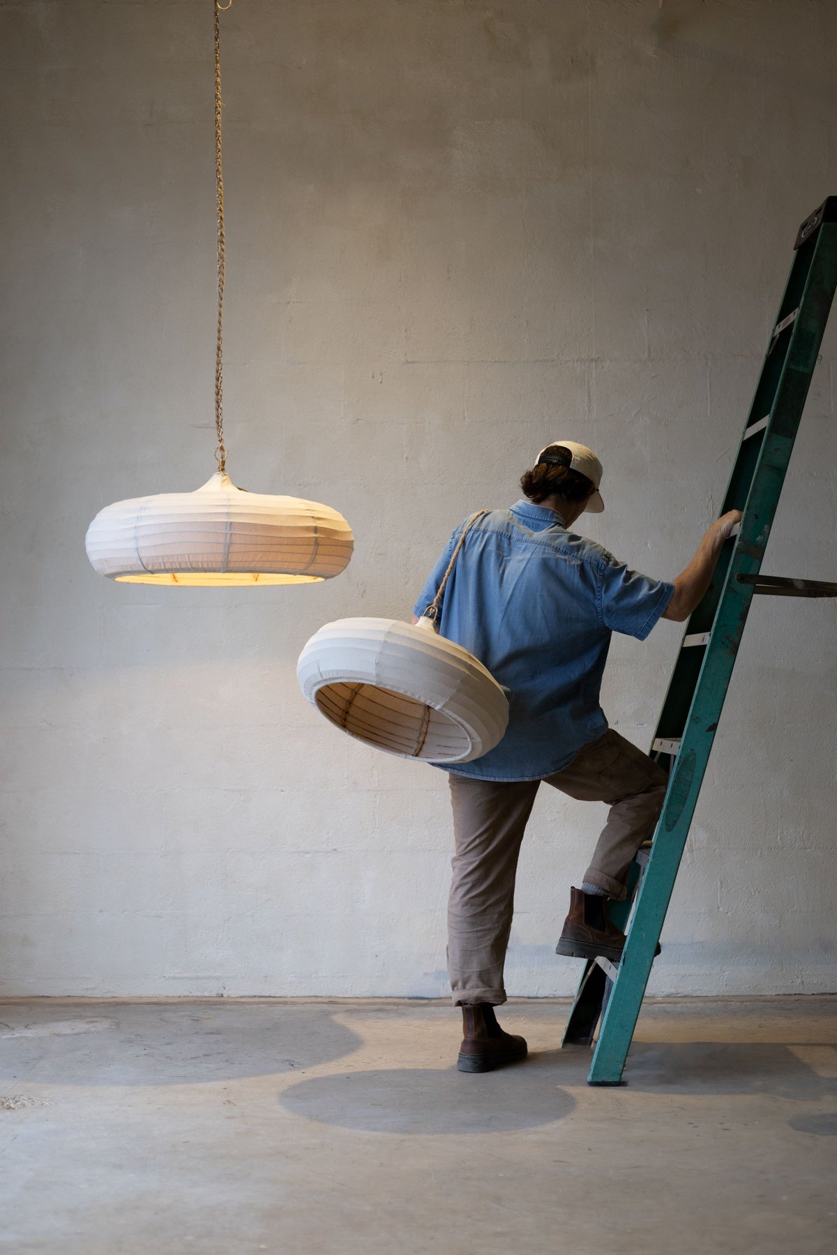 One teardrop pendant lights with off-white woven canvas hanging from ceiling while man steps of a green ladder to hang a second one.