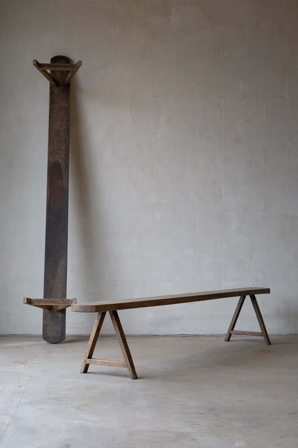 Two long, slim, wooden dining benches with simple A-frame. One bench is resting vertically on a neutral wall. Main image