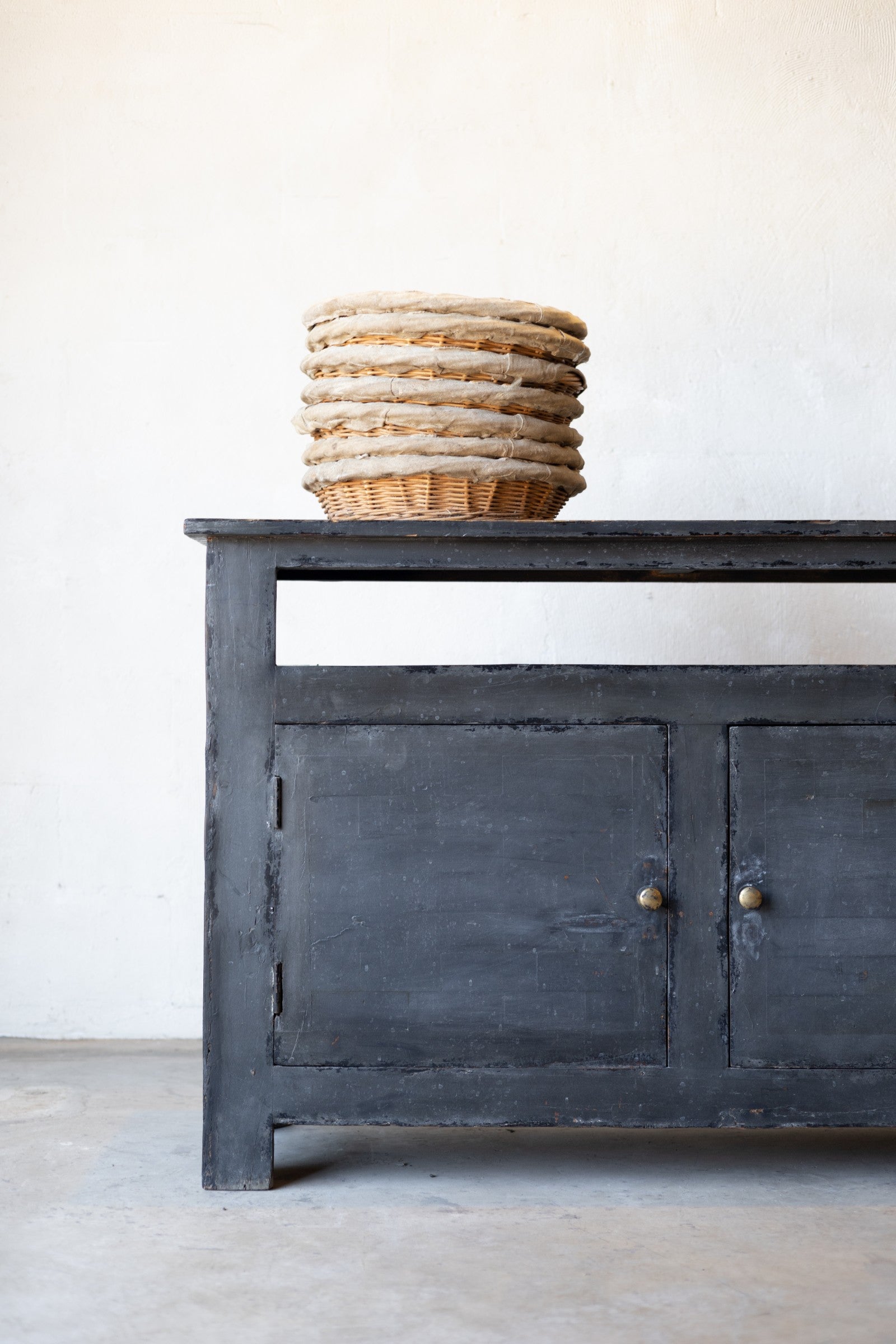 Dark wooden sideboard with a stack of woven baskets on a light gray floor against a white wall.