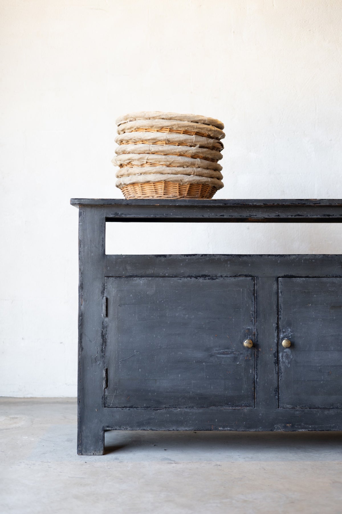 Dark wooden sideboard with a stack of woven baskets on a light gray floor against a white wall.