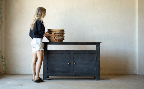 Woman arranging baskets on a black wooden console cabinet in a minimalistic room.