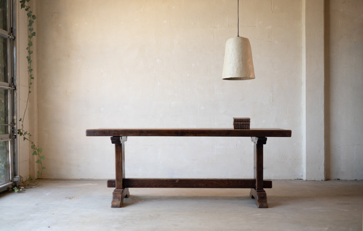 Side image of a long vintage oak dining table with a rustic design with books on the table and a white pendant light hanging over the table on a light background.