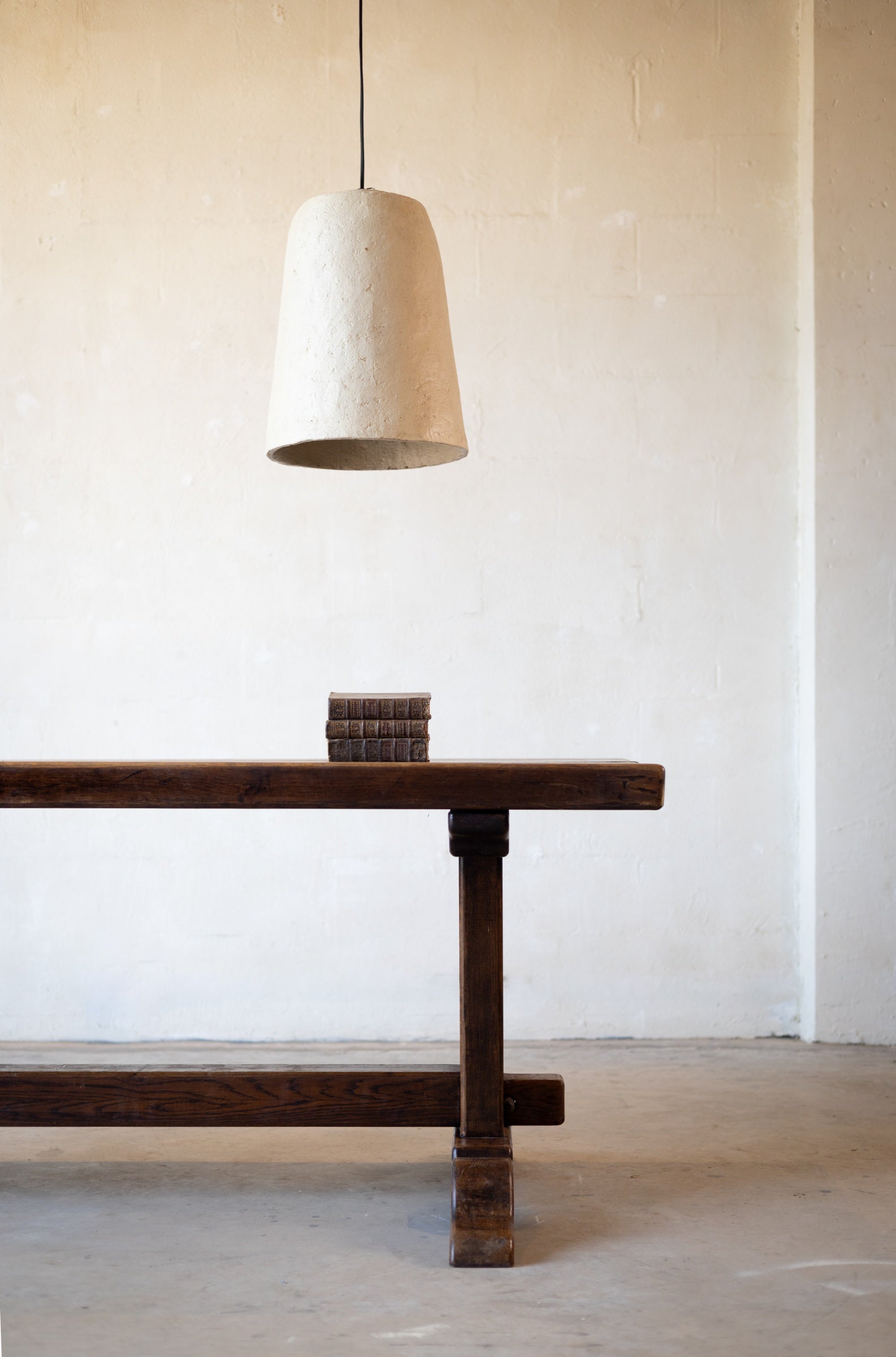 Cropped image of one end of a vintage oak dining table with a rustic design with books on the table and a white pendant light hanging over the table on a light background.