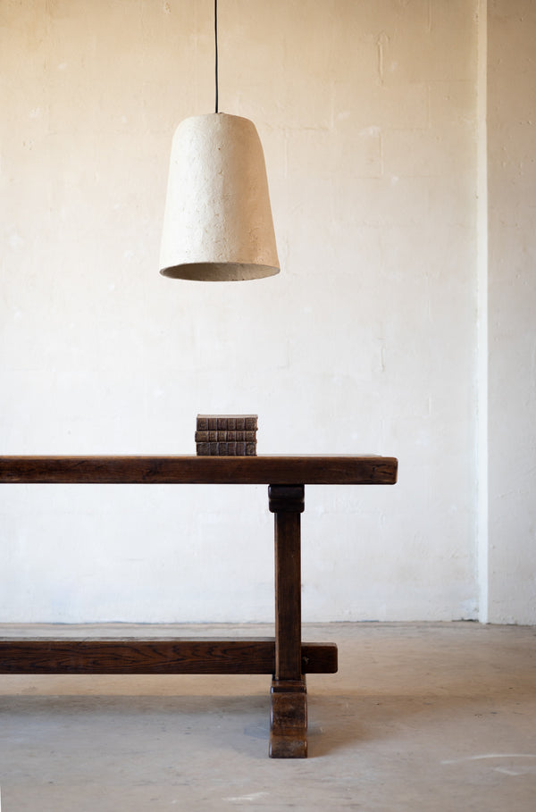 Cropped image of one end of a vintage oak dining table with a rustic design with books on the table and a white pendant light hanging over the table on a light background.