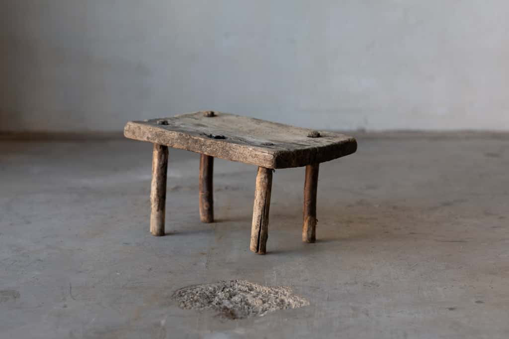 A small, wooden, rectangular workman's stool with rustic legs.