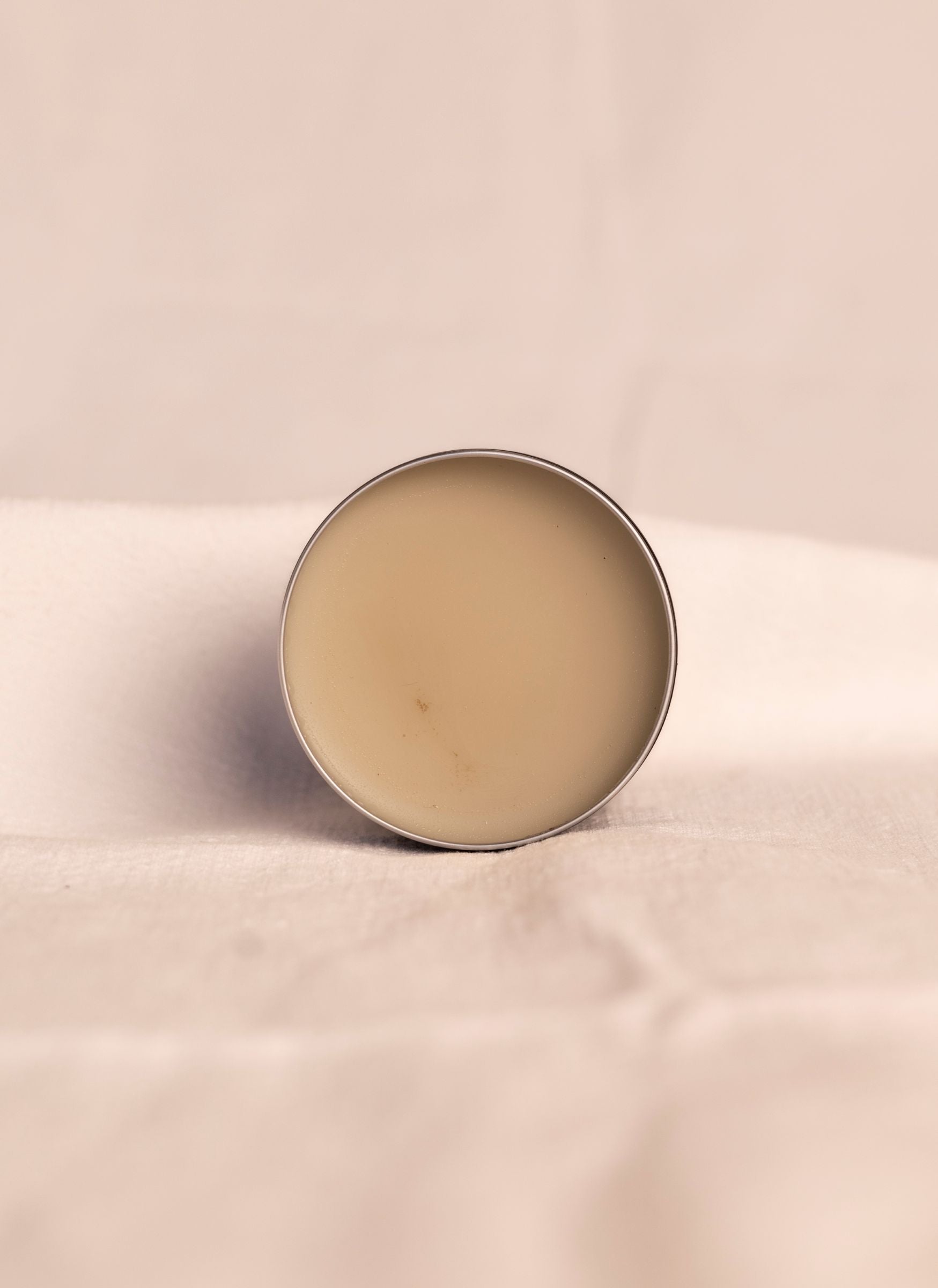 Open container of all natural ashe juniper moisturizing balm revealing beige product inside container against a soft white background.