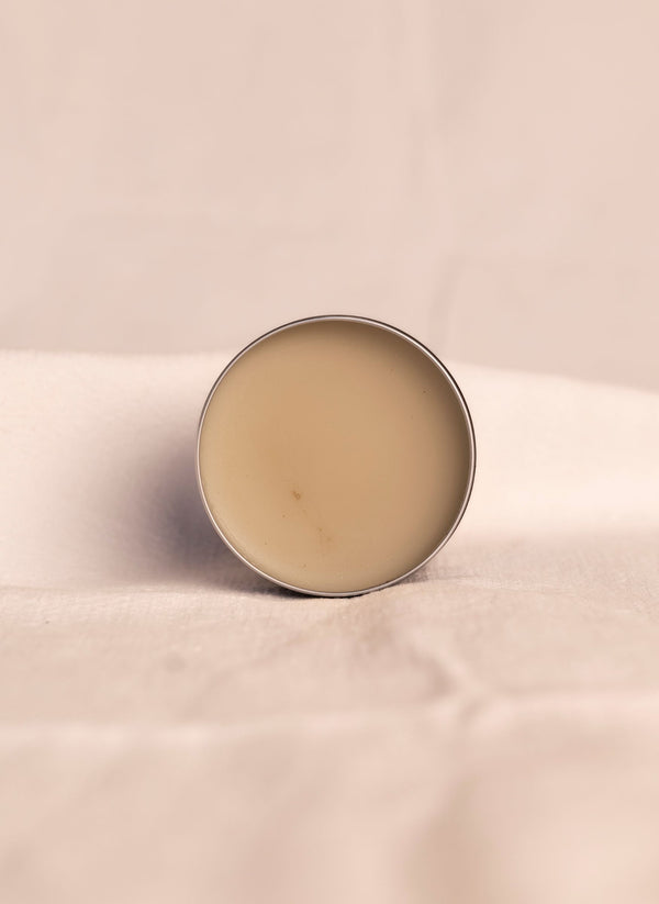 Open container of all natural ashe juniper moisturizing balm revealing beige product inside container against a soft white background.