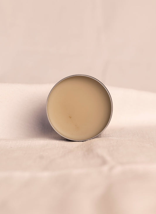 Open container of all natural ashe juniper moisturizing balm revealing beige product inside container against a soft white background.