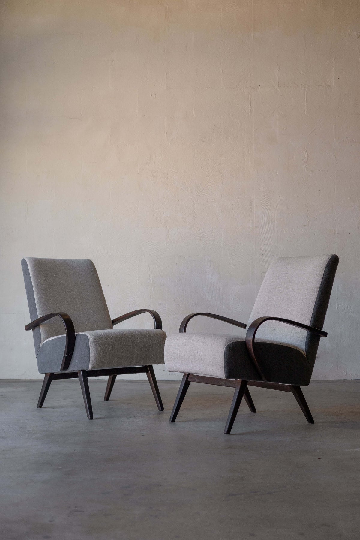 Pair of vintage Halabala lounge chairs with curved wooden armrests and wooden legs, featuring cream linen reupholstery on backrest and seat, and grey linen on chair's sides. Main image