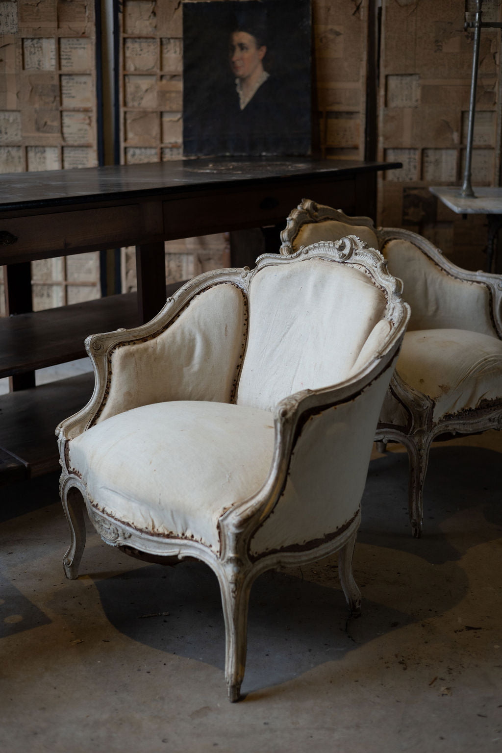 Upholstered off-white armchairs with intricate wooden carvings with dark wooden desk and portrait in the background. 