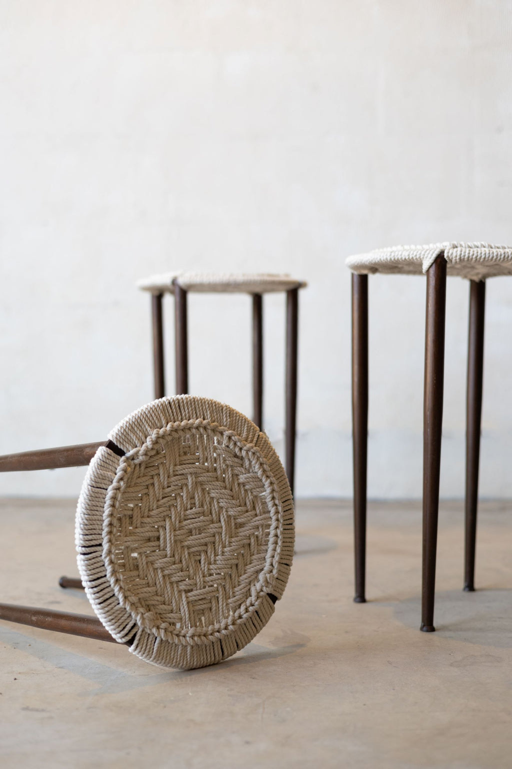 Iron stool tipped over revealing woven seat design and two chairs standing in background.