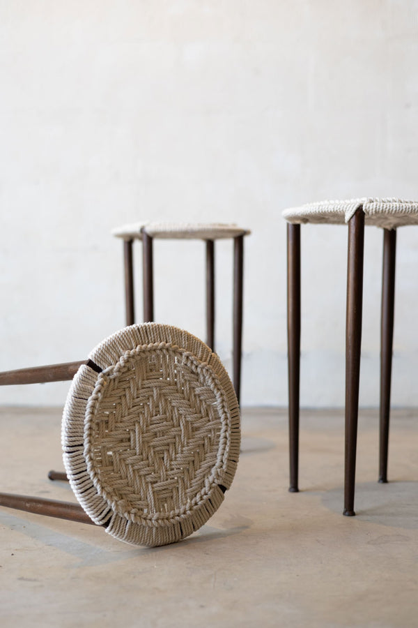 Iron stool tipped over revealing woven seat design and two chairs standing in background.