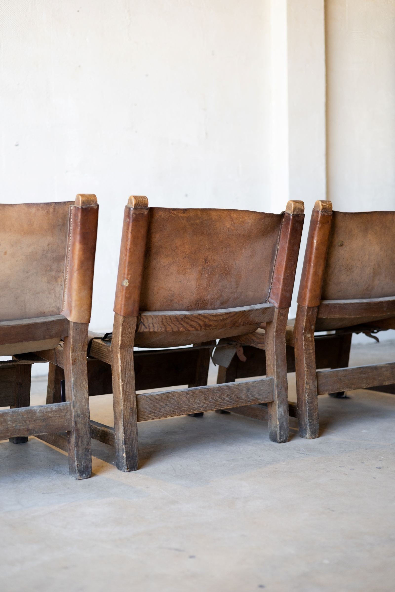 The backs of three vintage wooden and leather sling chairs with leather seats against a white wall.