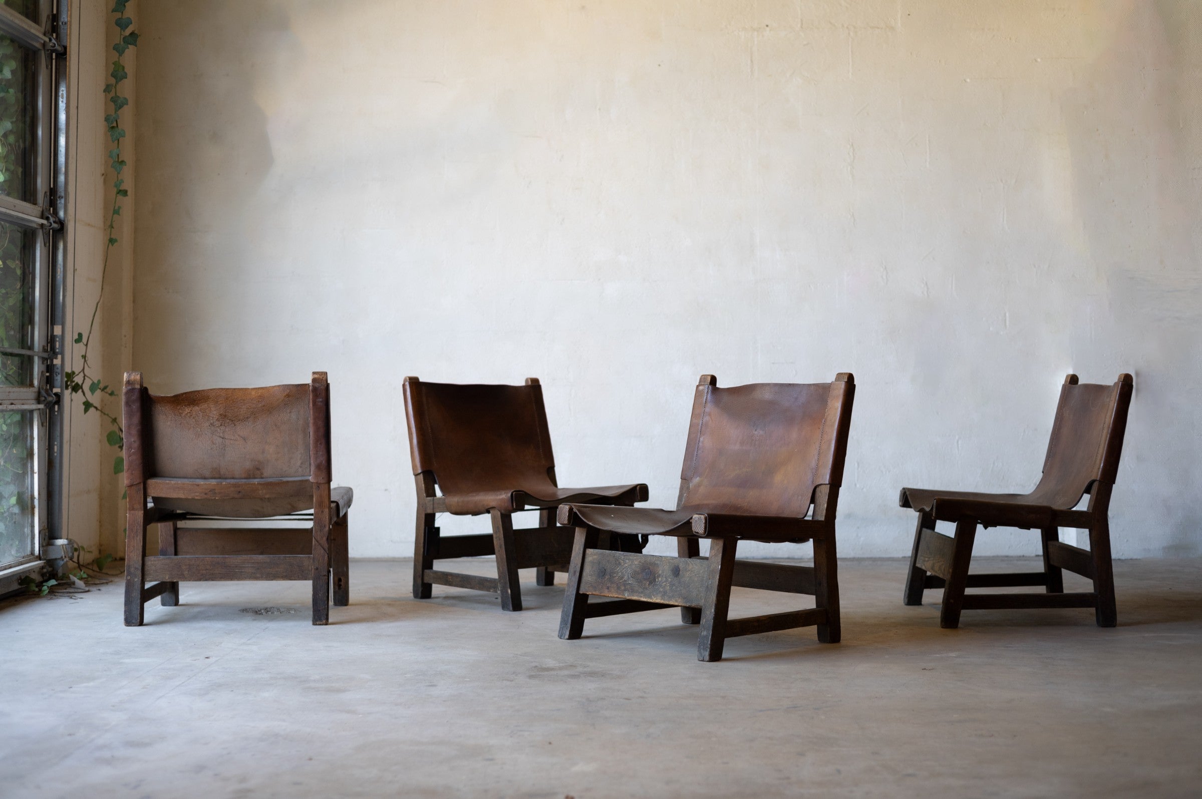 Four vintage brown wood and leather sling chairs arranged in a against a plain wall.