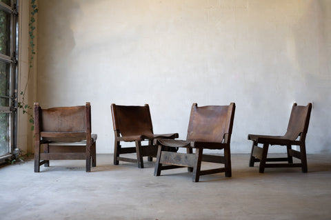 Four vintage brown wood and leather sling chairs arranged in a against a plain wall.