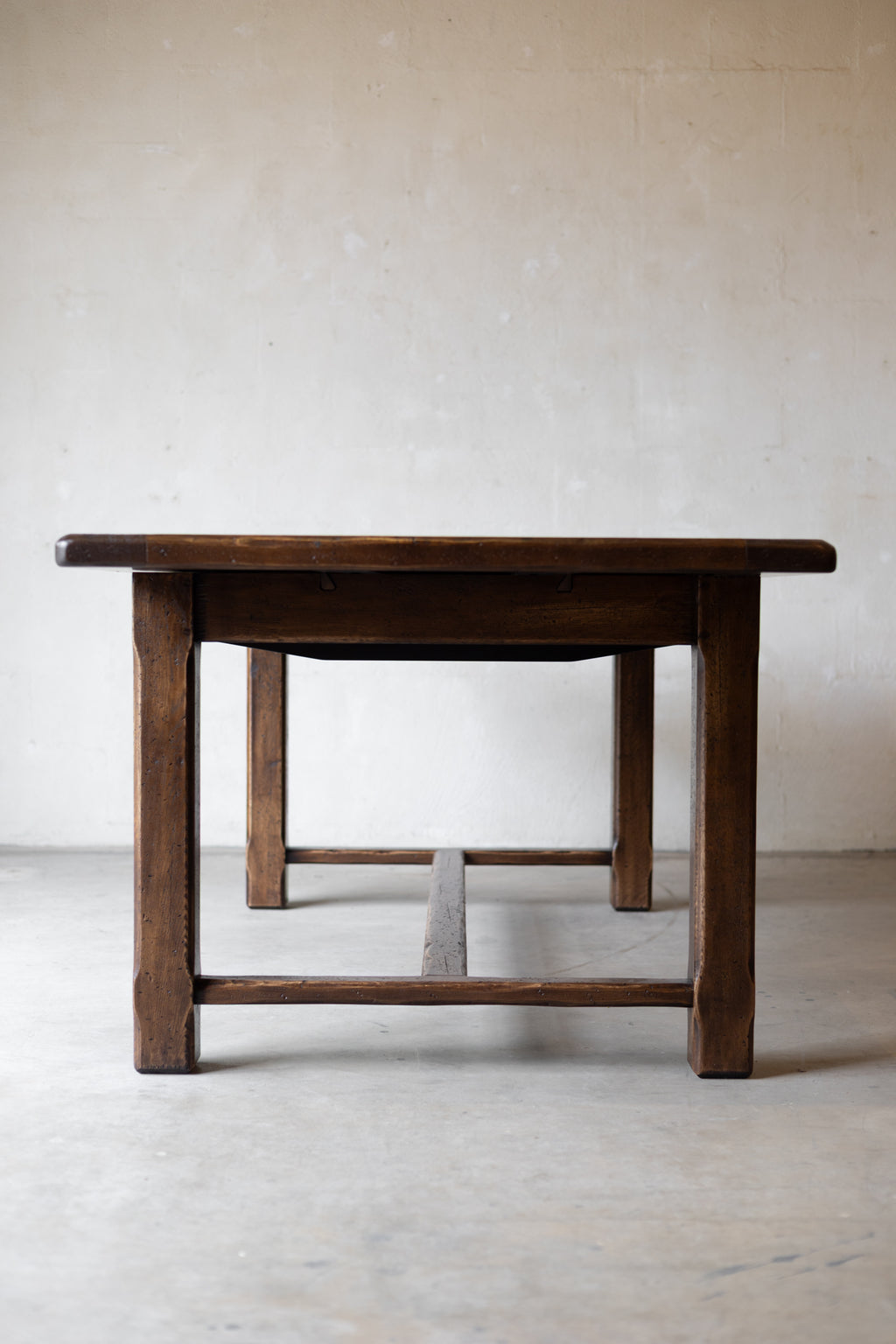 A dark brown wooden table with a thick rectangular top and four sturdy legs, shown from a low angle against a textured beige wall and concrete floor.