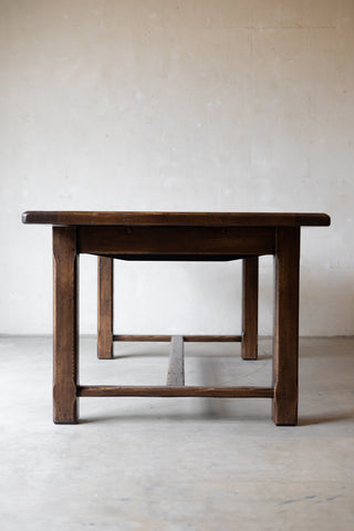 A dark brown wooden table with a thick rectangular top and four sturdy legs, shown from a low angle against a textured beige wall and concrete floor.