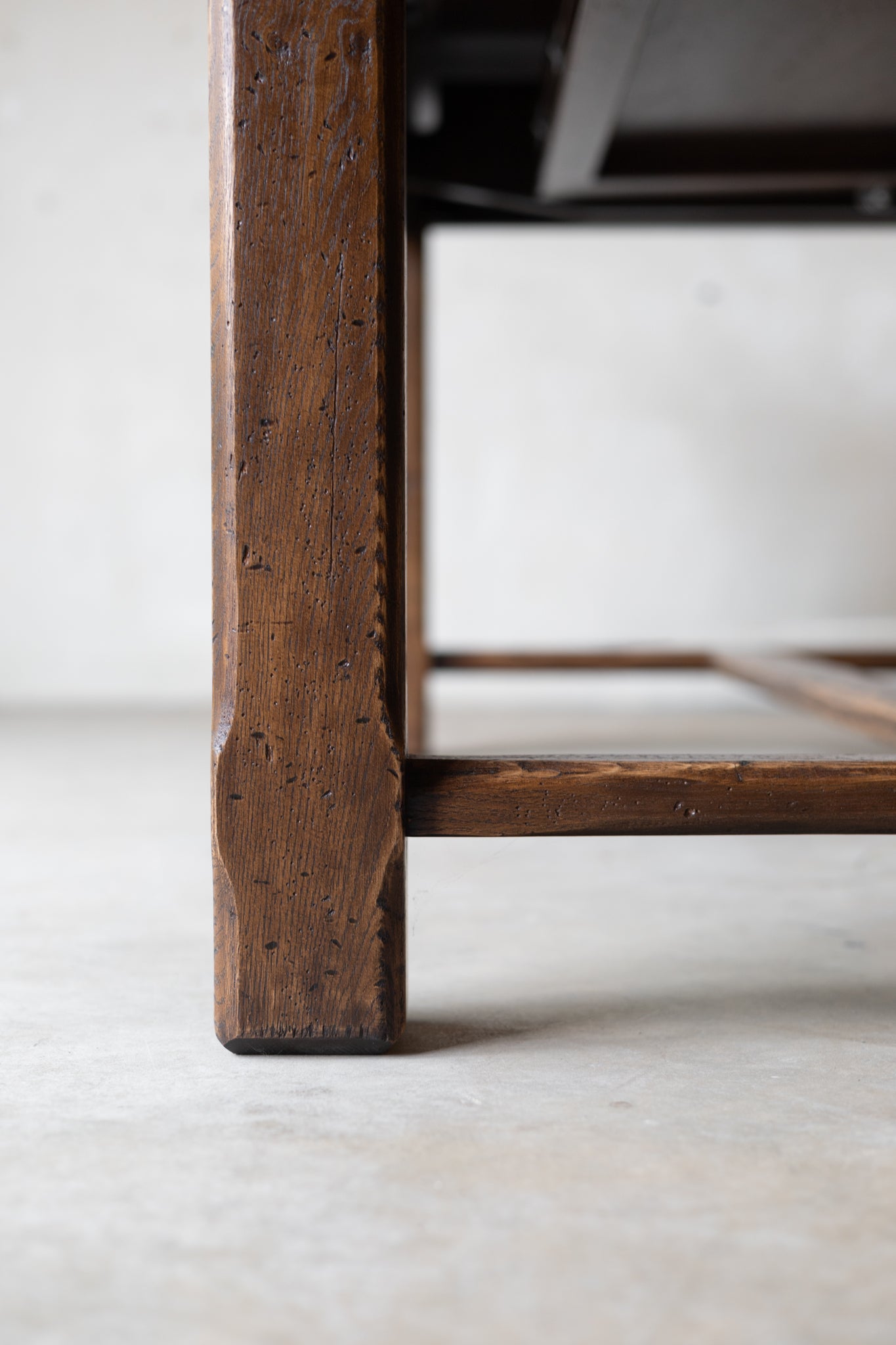 Close-up detail of a dark brown wooden table leg with visible grain and a footrest bar, set against a neutral light gray concrete floor and wall.