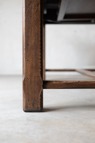 Close-up detail of a dark brown wooden table leg with visible grain and a footrest bar, set against a neutral light gray concrete floor and wall.