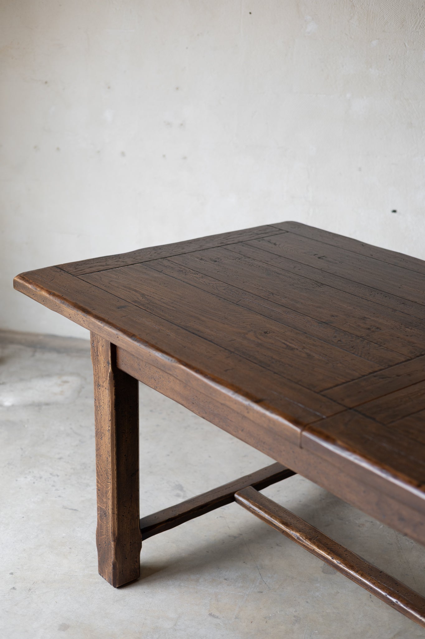 A dark brown wooden table with a live-edge natural top and a rustic crossbar support, photographed in a minimalist concrete room.