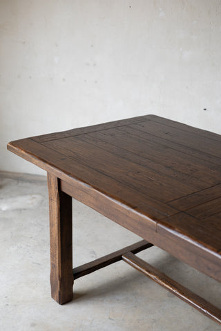 A dark brown wooden table with a live-edge natural top and a rustic crossbar support, photographed in a minimalist concrete room.