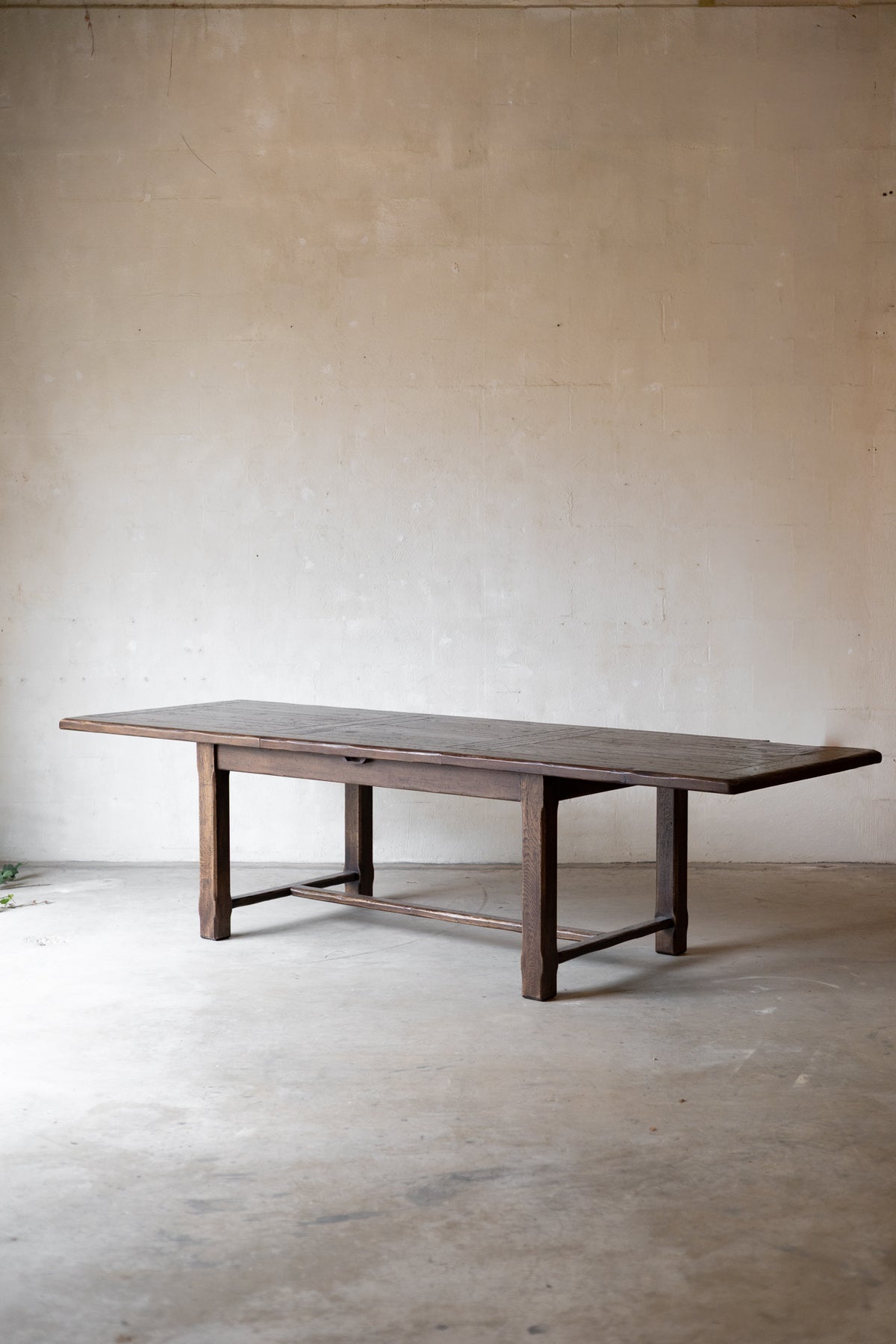 A dark brown wooden dining table with a rustic, weathered finish and a straight rectangular top, set against a minimalist concrete floor and textured plaster wall.