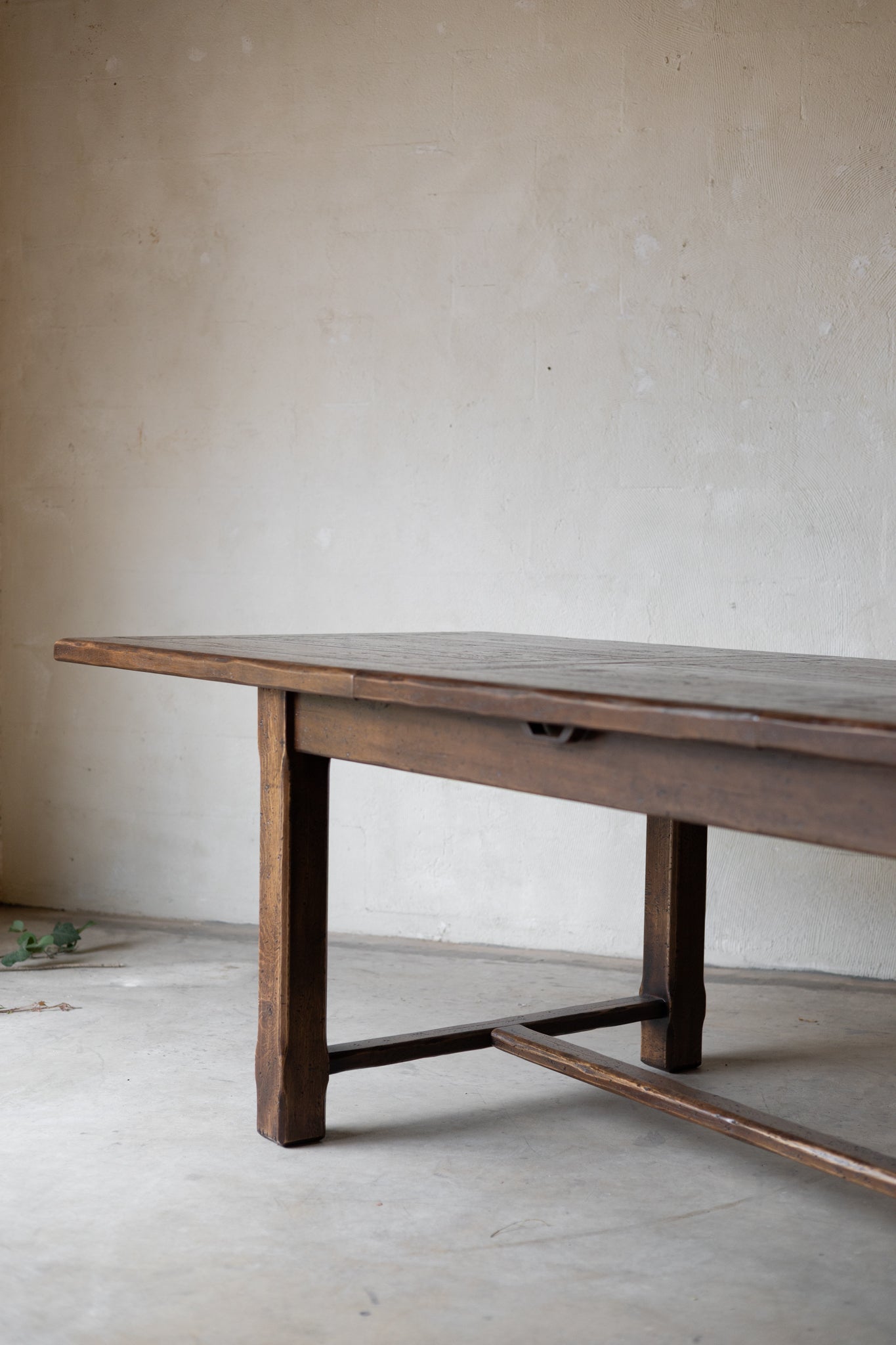 A rustic dark brown wooden table with a single drawer and stretcher base, photographed in a minimalist room with a textured plaster wall and concrete floor.