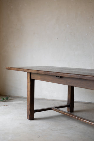 A rustic dark brown wooden table with a single drawer and stretcher base, photographed in a minimalist room with a textured plaster wall and concrete floor.