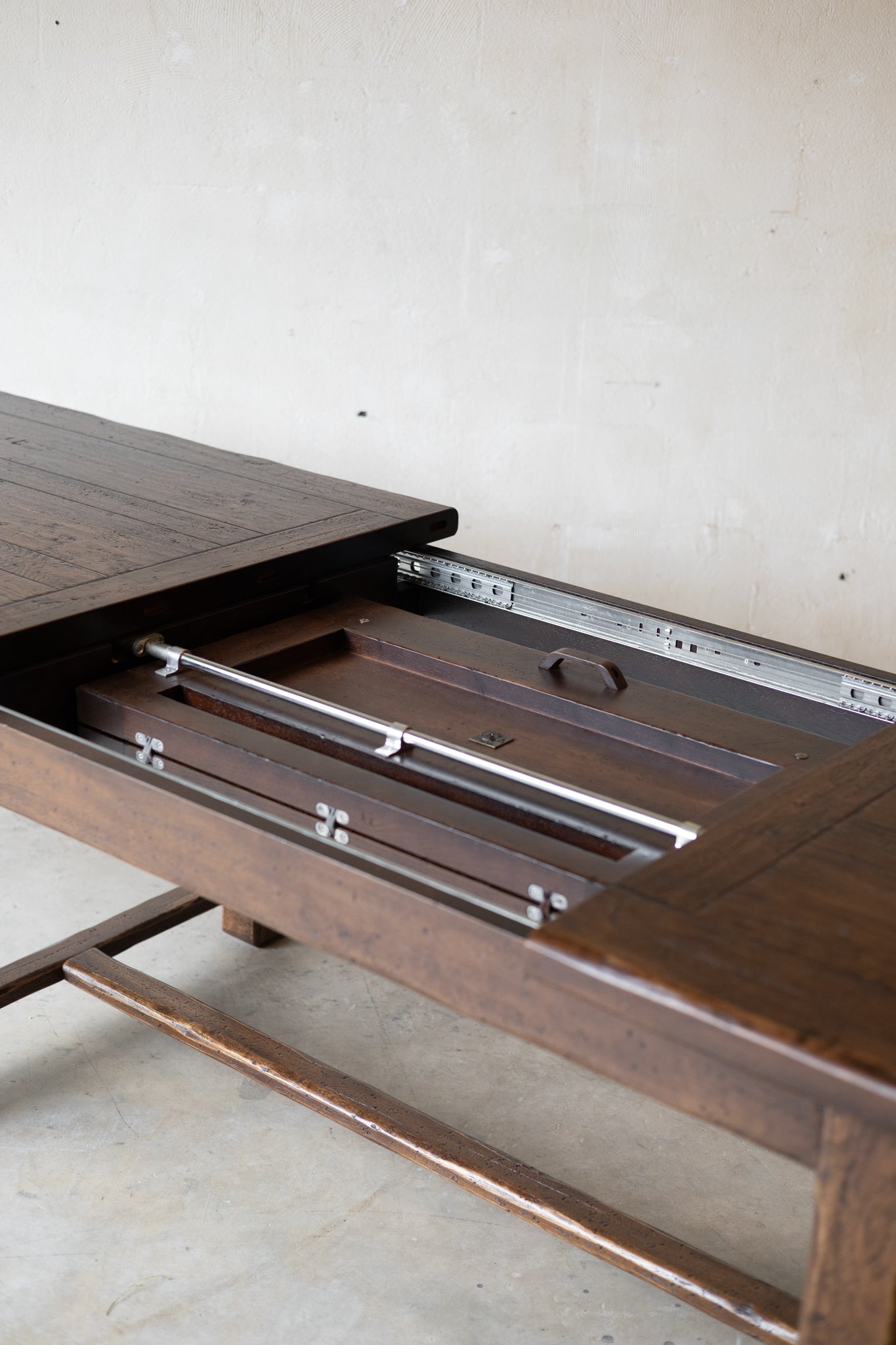 A close-up of a dark brown wooden table with a leaf pulled open, showing the metal leaf tracks and interior compartments against a plain light wall.