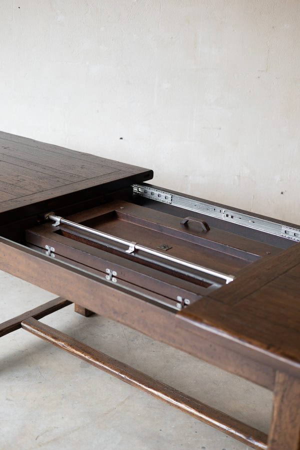 A close-up of a dark brown wooden table with a leaf pulled open, showing the metal leaf tracks and interior compartments against a plain light wall.