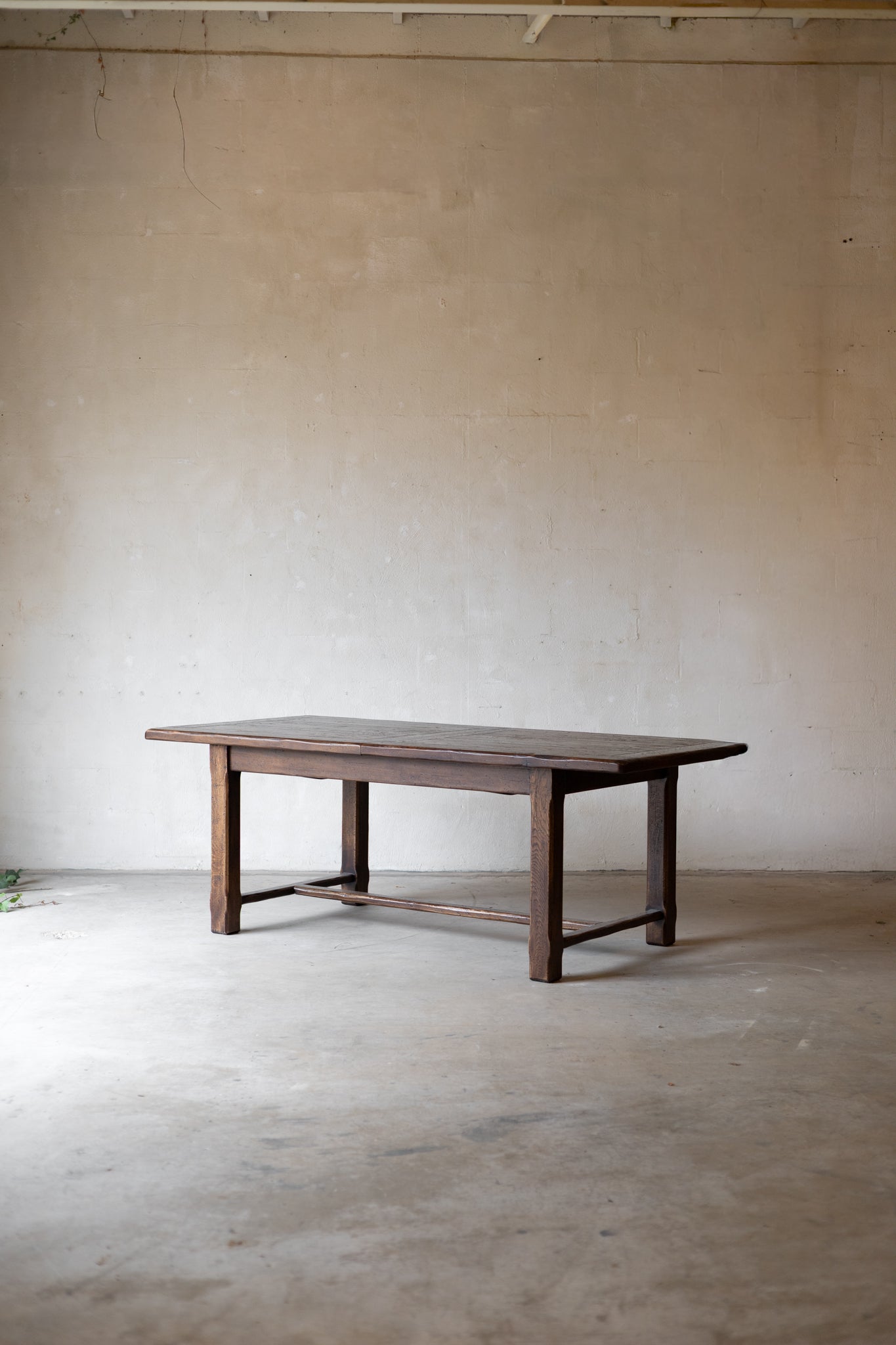A dark brown wooden dining table with a rectangular top and four legs, positioned in the center of a minimalist concrete room with a textured beige plaster wall.