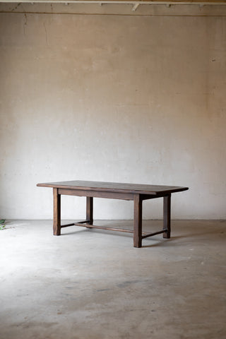 A dark brown wooden dining table with a rectangular top and four legs, positioned in the center of a minimalist concrete room with a textured beige plaster wall.