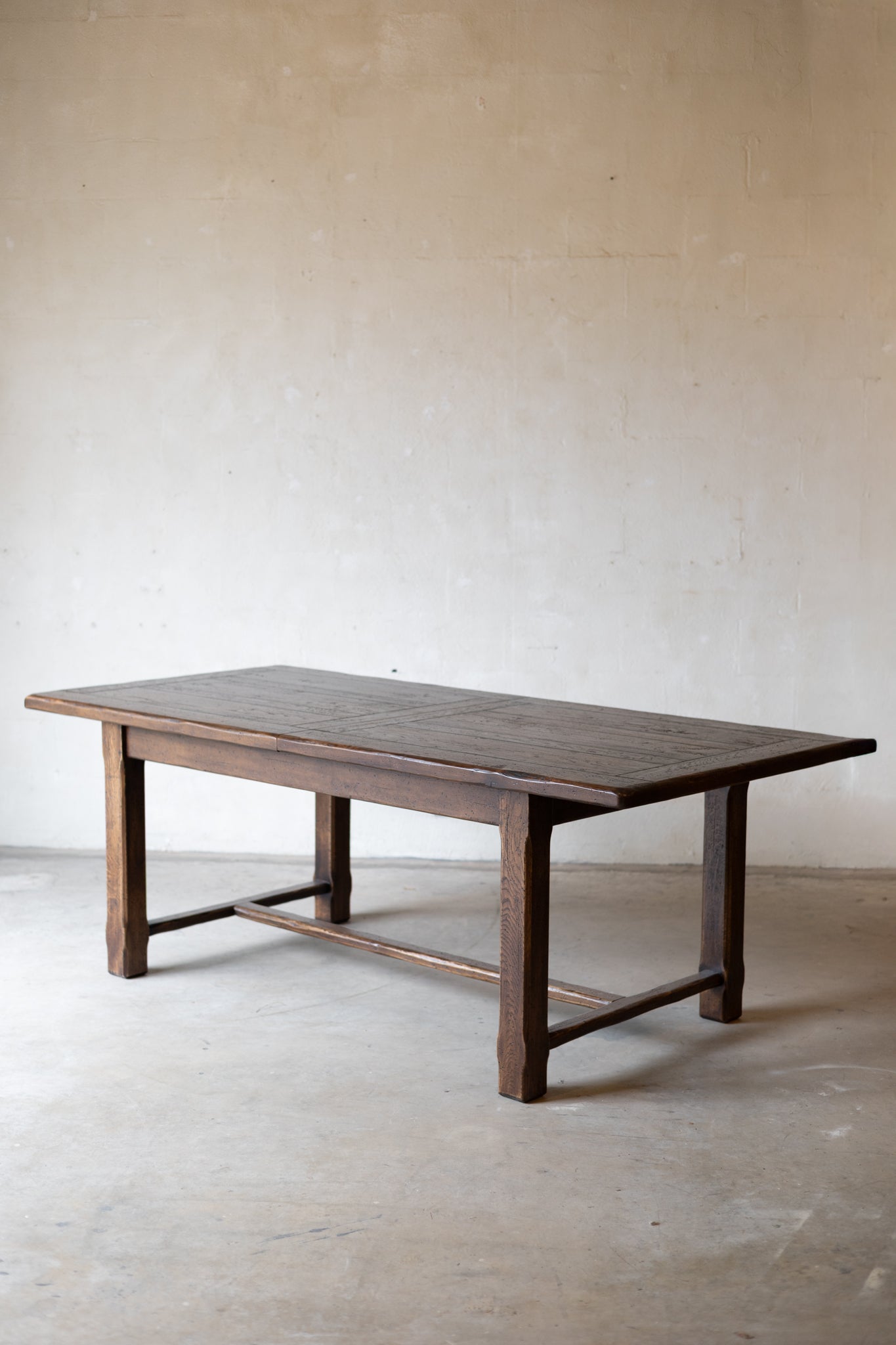 A rustic dark brown wooden table with a thick rectangular top and four sturdy legs, set against a neutral, textured beige plaster wall and concrete floor.