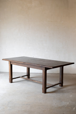 A rustic dark brown wooden table with a thick rectangular top and four sturdy legs, set against a neutral, textured beige plaster wall and concrete floor.
