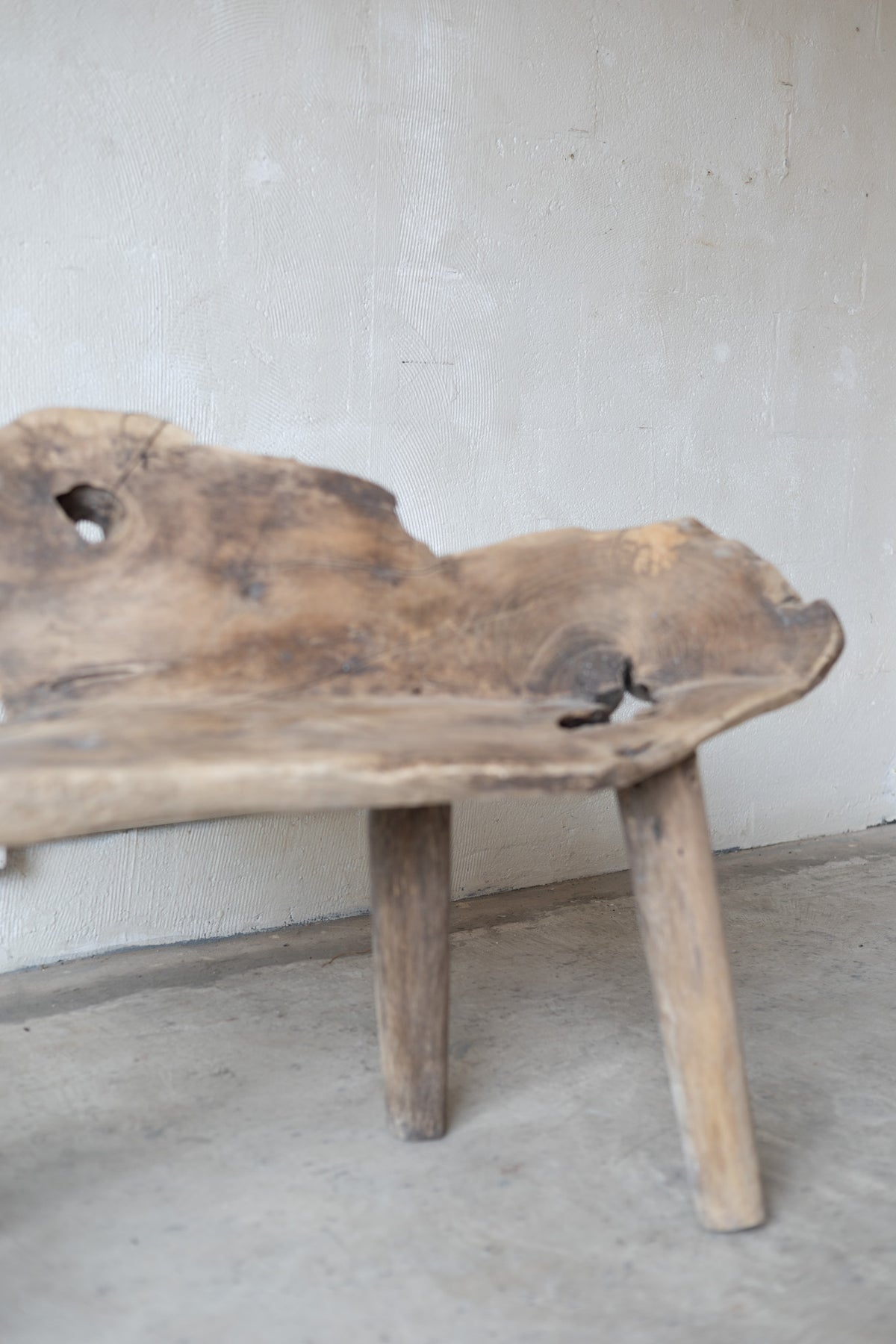A rustic live-edge wooden bench with a thick natural wood top and two sturdy legs, positioned against a textured light gray wall and concrete floor.