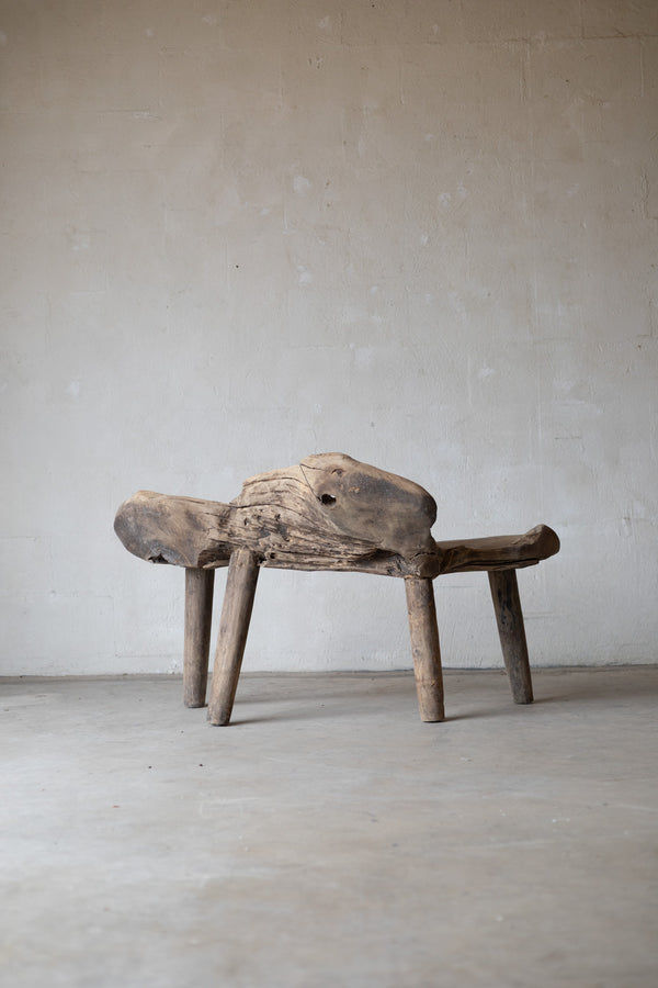 A rustic wooden bench made from a natural live-edge log, featuring a thick irregular top and four sturdy legs, set against a minimalist concrete wall and floor.