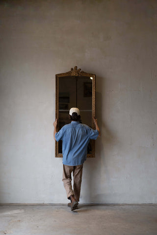 A man holding a full-length antique split glass mirror with an intricate gold frame.