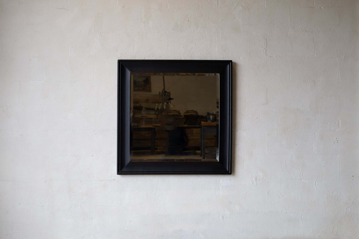 A square mirror with a black wooden frame with a beveled edge, hanging on a neutral wall.