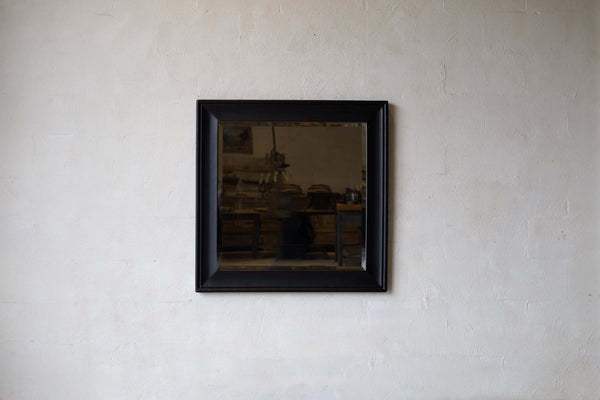 A square mirror with a black wooden frame with a beveled edge, hanging on a neutral wall.