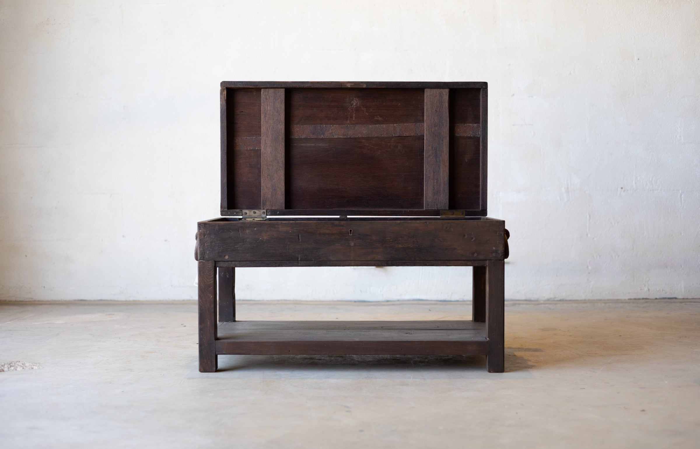 British colonial teak coffee table with open table-top compartment in revealing hidden storage in front a white textured wall.