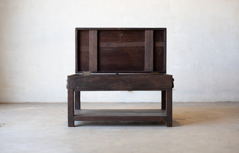 British colonial teak coffee table with open table-top compartment in revealing hidden storage in front a white textured wall.