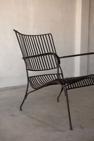 Side angle view of black iron lounge chair with vertical back slats, armrests, and curved seat.